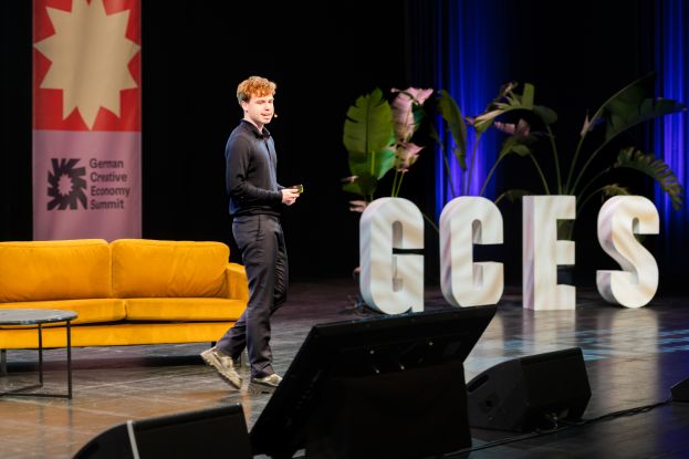 Excerpt from the German Creative Economy Summit. A stage with a speaker can be seen. In the background, there is a couch and the letters GCES., &copy; Jonas Walter