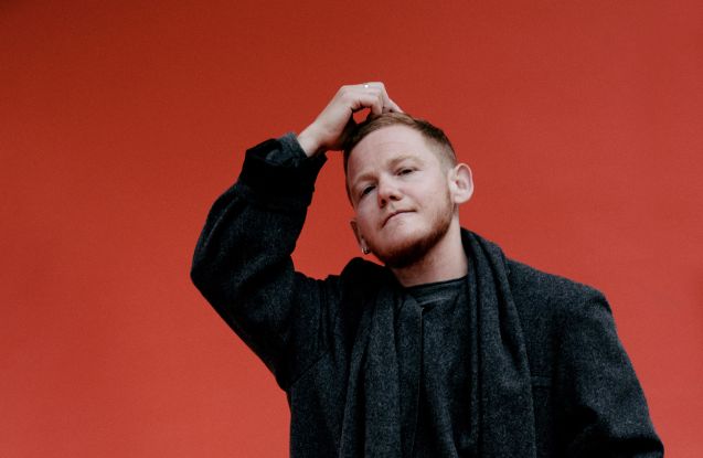 Kae Tempest stands dressed in black in front of a colorful wall with one hand on their head., &copy; Clara Nebeling