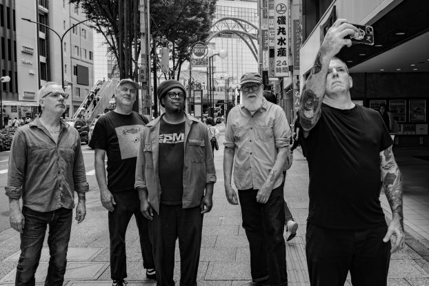 Five middle-aged male-looking people are standing together and looking up. One person with tattooed arms is holding up a cell phone and appears to be taking a selfie of the group. They are standing in the middle of a large Asian city., &copy; Yusuke Nagata